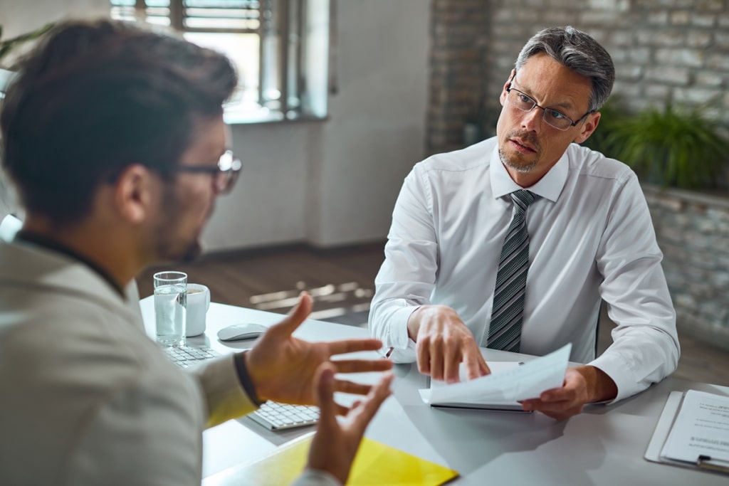 A imagem captura um momento intenso de conversa entre dois homens. O primeiro, vestido com uma camisa branca imaculada e uma gravata, aponta para um documento. O segundo homem, vestido com um blazer branco elegante, parece estar em pleno argumento, gesticulando com as mãos para enfatizar seus pontos.